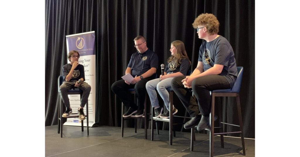 Jayson Lowe, CEO of Ascendant Financial, sits on stage with his children at an Ascendant Financial live event, beside a Becoming Your Own Banker banner